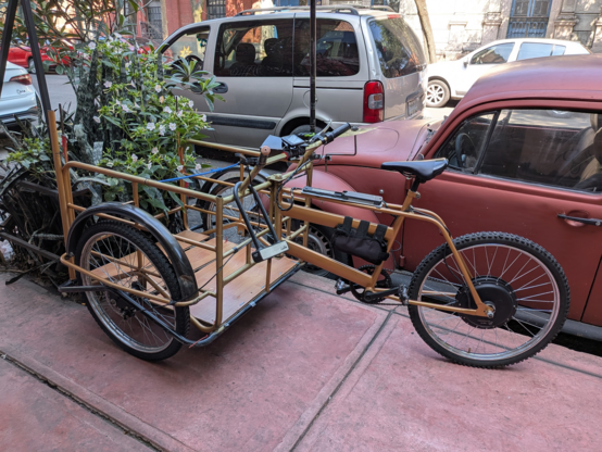 A heavy-duty Mexican cargo trike parked on the sidewalk near a street tree and an old Volkswagen Beetle in Mexico City. The trike's design is a classic, seen all across the country for decades. But this trike has an electric motor in its rear hub and a battery mounted in its main triangle. There is a small LCD display mounted on the handlebars.