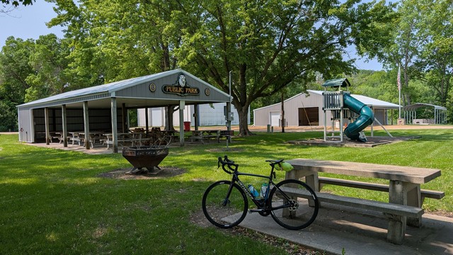 Blue and black Roubaix bicycle leaning against a concrete picnic table in a city park, shaded grass, partially open picnic shelter with tables, steel fire pit in the foreground, green plastic slide on the right and brown utility shed behind under sunny skies.