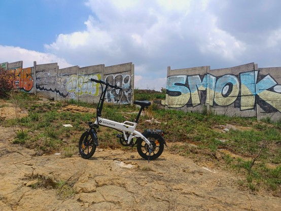 a photo of my ebike standing over stones of a hill in front of a wall with several graffitis.