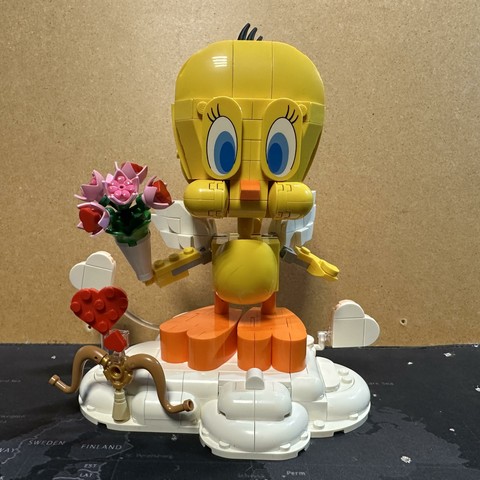 A Lego model of tweety the bird from Looney Tunes, holding a flower bouquet while standing on a cloud with hearts. Cupido’s bow and arrow are also included for some reason.
