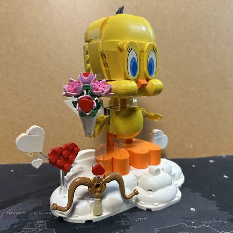 A Lego model of tweety the bird from Looney Tunes, holding a flower bouquet while standing on a cloud with hearts. Cupido’s bow and arrow are also included for some reason.