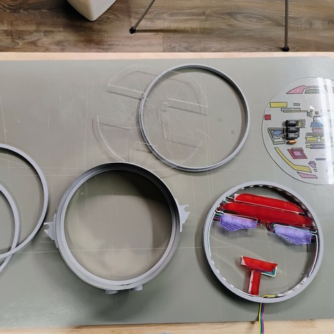 The components of Dr. Theo, laid out on a workbench.  The pieces are sitting on a grey piece of hard plastic.  From left to right, there are 2 spacer rings partially on top of each other, the main body, the piece of clear acrylic with pieces cut out, another spacer, the segment with all of the lights attached, and the segment with the colored shapes and fake electronics attached.