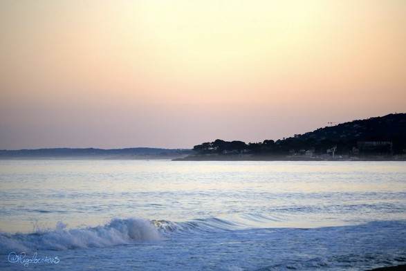 Photo du bord de mer peu après le coucher du soleil. La mer a pris une teinte bleue très pâle, presque blanche. On voit un petit rouleau de vague se briser et de l’écume au premier plan. À l’horizon, un bout de colline se découpe en noir sur le fond de ciel rose pâle. Le ciel est un dégradé de gris, rose orangé et jaune.