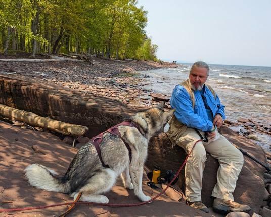 Hiker Jeff sits on a long flat rock rising from a rocky shoreline, eating nut clusters out of a ziplock bag. He's holding it slightly to the side, out of reach of hungry Tonttu, who is sitting very attentively at his side, head and nose pointed directly at the bag.