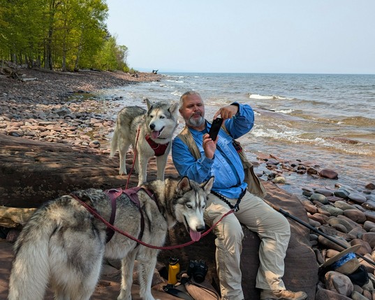 Still sitting on the rock where snacks were eaten, Jeff leans back towards Zeena, who is now standing on the rock beside him, to take a selfie with the two of them together, though Zeena is distracted by something to the left. Tonttu stands in front, looking back at the camera with a somewhat mischievous look on his face. Lake superior ripples in the background, along with a shoreline that recedes towards the horizon, and the sky is a uniform grey-blue of haze.