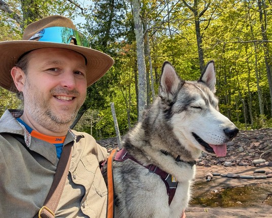 Dale and Tonttu pose for a selfie on the shoreline. The camera points back towards the birch and aspen trees glowing yellow green with backlit sun behind them. Dale is smiling at the camera, while Tonttu has his eyes squinted closed, with a small bit of tongue hanging out of his open-smile muzzle.