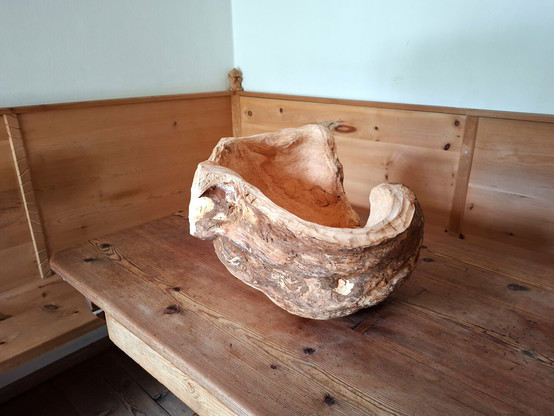 A large, organically shaped wooden sculpture or natural woodform rests on a larch wood table in the corner of a room. The room features warm-toned, knotty arolla pine paneling and built-in bench seating. The object appears to be carved from a single, solid piece of wood, with flowing curves and hollowed areas that resemble an abstract bowl or natural seat. The surface showcases a mix of smooth, pale sections and rough, textured bark-like areas, emphasizing its raw and sculptural qualities. A white curtain hangs above the paneling, and soft natural light enhances the calm, rustic atmosphere. In the corner on the end moulding of the pine panelling is a small hand-carved figure with a mushroom head.