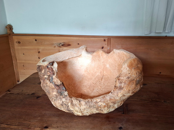 A large, organically shaped wooden sculpture or natural woodform rests on a larch wood table in the corner of a room. The room features warm-toned, knotty arolla pine paneling and built-in bench seating. The object appears to be carved from a single, solid piece of wood, with flowing curves and hollowed areas that resemble an abstract bowl or natural seat. The surface showcases a mix of smooth, pale sections and rough, textured bark-like areas, emphasizing its raw and sculptural qualities. A white curtain hangs above the paneling, and soft natural light enhances the calm, rustic atmosphere.
in the corner on the end moulding of the pine panelling is a small hand-carved figure with a mushroom head