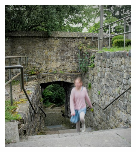 A blurred, slow-shutter image of a child running up some steps with a stone arch in the background. The child is wearing a pink jumper and blue jeans skirt and has long hair.