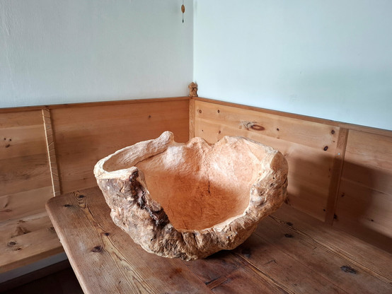 A large, organically shaped wooden sculpture or natural woodform rests on a larch wood table in the corner of a room. The room features warm-toned, knotty arolla pine paneling and built-in bench seating. The object appears to be carved from a single, solid piece of wood, with flowing curves and hollowed areas that resemble an abstract bowl or natural seat. The surface showcases a mix of smooth, pale sections and rough, textured bark-like areas, emphasizing its raw and sculptural qualities.