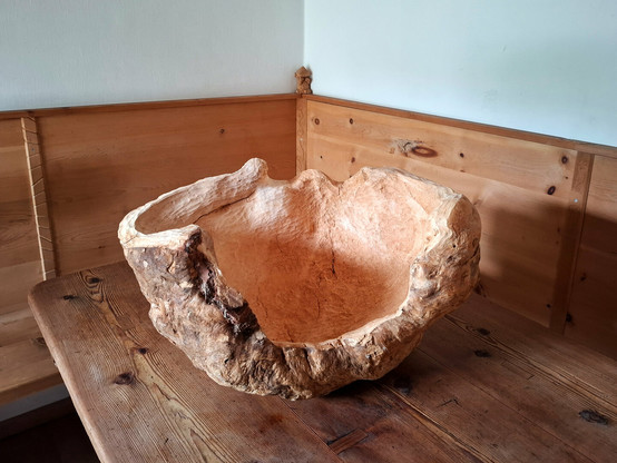 A large, organically shaped wooden sculpture or natural woodform rests on a larch wood table in the corner of a room. The room features warm-toned, knotty arolla pine paneling and built-in bench seating. The object appears to be carved from a single, solid piece of wood, with flowing curves and hollowed areas that resemble an abstract bowl or natural seat. The surface showcases a mix of smooth, pale sections and rough, textured bark-like areas, emphasizing its raw and sculptural qualities.