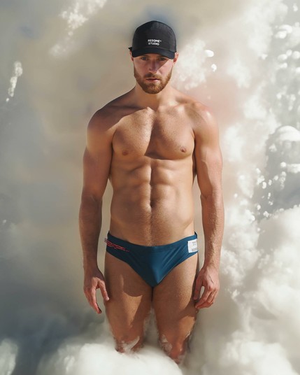 A muscular white guy wearing speedos is standing in front of a big wave of bubbles