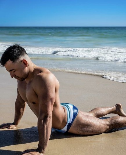 A muscular guy wearing speedos is doing yoga on a beach