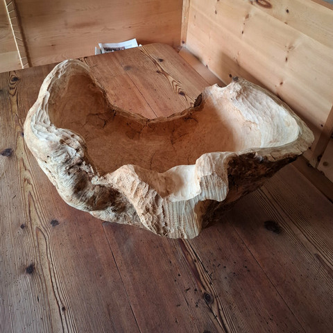 A large, organically shaped natural woodform rests on a larch wood table. The room features warm-toned, knotty arolla pine paneling and built-in bench seating. The object appears to be carved from a single, solid piece of wood, with flowing curves and hollowed areas that resemble an abstract bowl or natural seat. The surface showcases a mix of smooth, pale sections and rough, textured bark-like areas, emphasizing its raw and sculptural qualities.