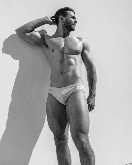 A black and white photo of a muscular white man wearing white speedos posing in sunshine