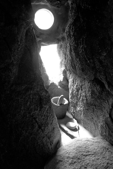 The image depicts a narrow, rocky passage with a small circular window at the top, allowing light to enter. The walls of the passage are rough and textured, with visible cracks and crevices. The light from the window creates a stark contrast between the illuminated area and the surrounding darkness. In the center of the passage, there is a stone mortar and pestle, along with a round stone, placed on a flat surface. The floor of the passage is uneven and covered with a layer of dust or fine particles. The overall scene is monochromatic, emphasizing the textures and shapes within the rocky environment.

Provided by @altbot, generated privately and locally using Ovis2-8B