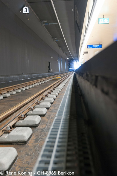 Das Bild zeigt einen unterirdischen Bahnsteig mit einer perspektivischen Ansicht der Gleise, die sich in die Ferne erstrecken. Das Gleis ist mit der Nummer "3" auf einem Schild darüber gekennzeichnet.
Die Gleise bestehen aus Metallschienen, die auf Betonschwellen gelagert sind, wobei auf dem Metall ein leichter Rost sichtbar ist. Der Bahnsteig wird auf der rechten Seite von einer niedrigen Mauer begrenzt, und an der Decke befinden sich Beleuchtungskörper und ein Hinweisschild mit Richtungsangaben.
Die Beleuchtung ist hell, mit einer starken Lichtquelle am Ende des Bahnsteigs, der Tunnelöffnung. Die Wände sind dunkel, und die Gesamtatmosphäre ist sauber und modern.