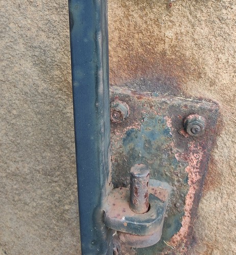 A rusty lower hinge bracket  is bolted onto brownish sandstone, supporting an old  wrought iron gate painted faded blue. The bracket looks like a face peering off to the left.
