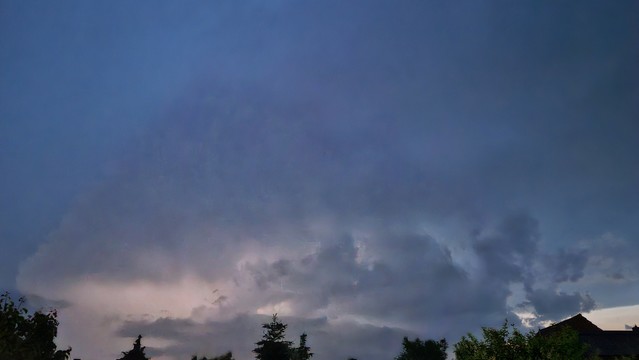 Photo of the sky covered in dark blue clouds. There's a dome like structure of thunderstorm clouds rising high into the sky. The internal glow of lightening  illuminates the clouds from the inside and shows the contrast between the cloud itself and the surrounding smaller clouds and the cloud cover far above. A small bright sliver of actual sky on the lower right side makes it clear that it would still be daylight without the thunderstorm, but the clouds have darkened everything till it appears like night fall.