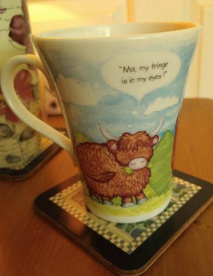 Mug of tea on a bedside cabinet with a Highland cow in a field, a speech bubble above it's head, saying 'Ma my fringe is in my eyes '