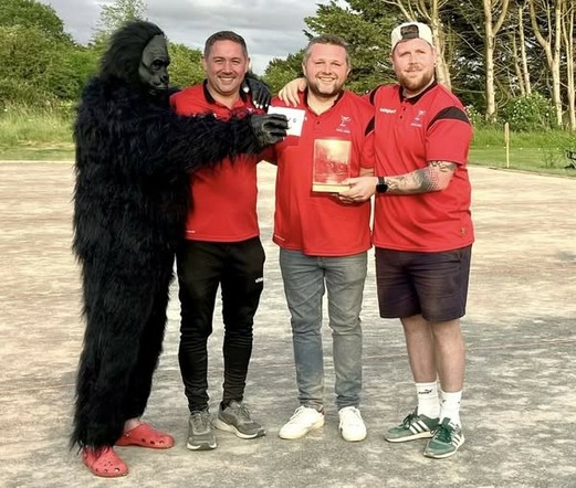 The image shows four individuals standing outdoors on a paved area with trees in the background. The person on the left is dressed in a black gorilla costume with red Crocs on their feet. The other three individuals are wearing matching red polo shirts with black accents, suggesting they are part of the same team or organization. The person in the middle is holding a plaque with a red and gold design, while the person on the right is holding a white card. The person in the gorilla costume is handing the card to the person in the middle. All four individuals are smiling and appear to be in a celebratory mood.

From @altbot