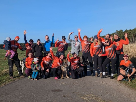 Eine Gruppe von knapp 20 Menschen, viele davon in orange-farbener Kleidung des Vereins Hauptstadt Canicross, lächeln und winken in die Kamera. Das Foto zeigt einen kleinen Teil unserer Mitglieder, die bei einem Schlittenhunderennen in Brandenburg gestartet sind.
