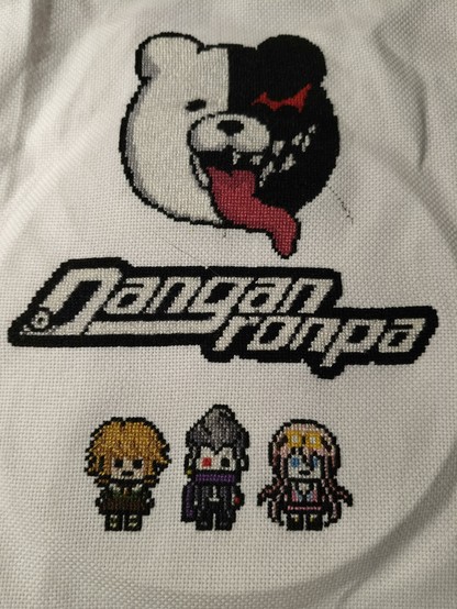 a cross stitch on white hardanger weave. At the top is the head of Monokuma with his tongue sticking out. in the middle is the Danganronpa logo in black and white. at the bottom is one character from each game, Chihiro from 1, Gundham from 2, and Miu from v3.