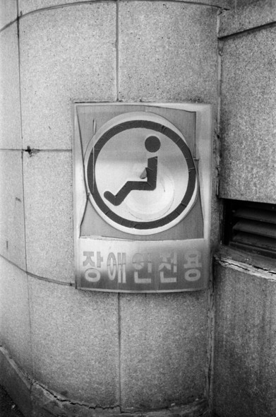 장애인 전용 주차구역을 알리는 간판이 기둥에 붙어있다. 얇은 철판에 휠체어 픽토그램이 붙어있는 형태인데 픽토그램의 바퀴 부분이 떨어져나가 사람이 허공에 앉아있는 모양이 되었다.