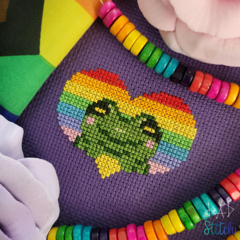 A cross stitch project staged with a Progressive Pride flag, some fake flowers, and rainbow colored beads. The project is stitched on purple fabric and features a rainbow heart with a smiling frog snug inside the heart's border. BAD Stitch.