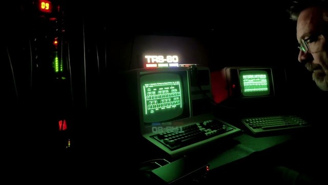 Image from Downpoly, bearded man with glasses staring into the green glowing CRT screens of two Tandy TRS-80 computers, TRS-80 letters glowing in the center screen. Live music from the TRS-80 Model 4s, Orchestra-90 and MIDI/80.
