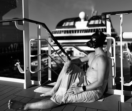 A black/white monochrome picture showing a hooded human dog relaxing on a cruise ship sundeck. He wears a black/camo pup hood with pointed ears and a white nose ring.