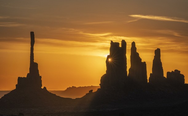 The rising sun creates a sunstar through a hole in a sandstone pillar rock formation in the desert. The red desert sand contributes to spectacular sunrise of orange and yellow.