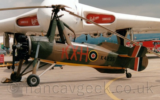 Side view of a 2 seat, single-engined gyrocopter parked in the static display park at an airshow, facing to the left.
The plane is mostly covered in an olive green and brown camouflage scheme, with a bright yellow belly.
The black serial "K4235" is on the rear fuselage, just aft of a blue, white, and red roundel outlined in yellow, itself just aft of  the red letters "KX-H" under the rear cockpit.
The 3 tails each have a large red, white, and dark blue fin flash.
There are 4 supports around the open front cockpit, heading upwards, with a helicopter-style rotor at the top.
A large white, delta-winged jet-engined military bomber aircraft dominates the background, with red covers with white text "Vulcan" and "XM603" in the engine inlets in the wing roots.
Grey sky fills the rest of the frame.
