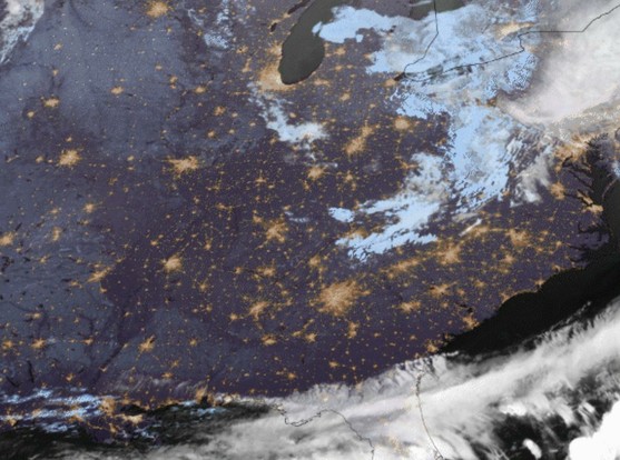 The image is a satellite view of the northeastern United States at night. The dark blue areas represent the night sky, while the yellow and orange lights indicate urban areas with artificial lighting. The brightest clusters of lights are concentrated in the northeastern part of the image, suggesting major cities like New York City and Philadelphia. The lights are more sparse in the central and western parts of the image, indicating less urban development. Clouds are visible as white and light blue patches, with a significant cloud formation in the upper right corner. The coastline of the Atlantic Ocean is visible along the eastern edge of the image, with the state of New Jersey and parts of New York and Pennsylvania identifiable by their shapes