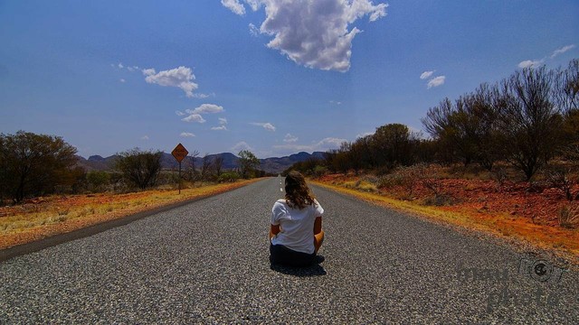 A person with long hair, wearing a white shirt, sits in the middle of an empty paved road. Surrounding landscape features dry vegetation, scattered trees, and distant mountains. A single cloud hovers in a blue sky. A road sign is visible on the left side.