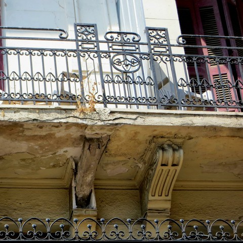 An upward view of the balcony of a classical-style building. Two supporting cantilevers are visible—one of them damaged and revealing a reinforced concrete beam inside.