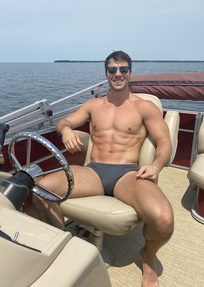 A muscular white man wearing speedos smiling at the camera while manspreading on a boat