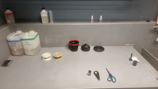 A table with equipment laid out for developing black & white films. Several bottles of chemicals; a developing tank and its constituent parts; two reels; scissors; the bottle opener thing for opening the film cannister.
