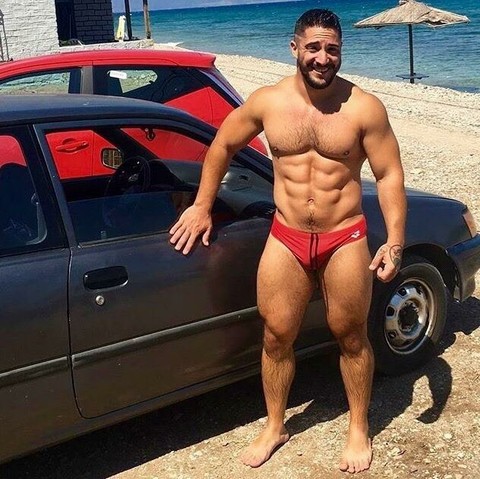 A muscular man in red speedos touching an old car and smiling at the camera