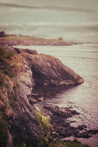 A dramatic coastal scene featuring a rugged, rocky cliff face meeting the calm waters of the sea under a cloudy sky. The cliffs show various textures and colours, with some green moss clinging to the darker rock. In the distance, the Wicklow coastline stretches further, with a headland and hints of a town or village on the horizon. The overall mood is slightly muted and contemplative, with a vintage aesthetic.