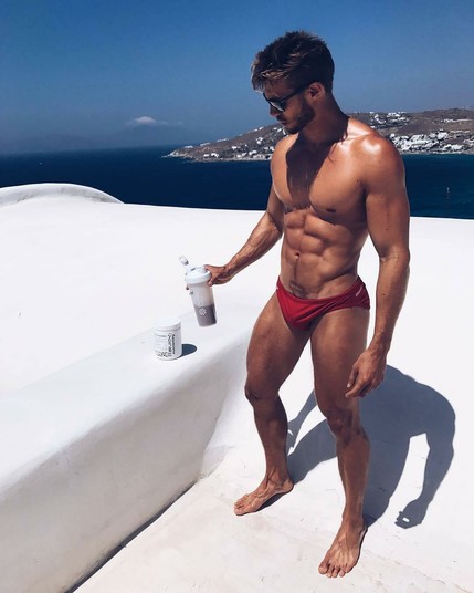 A muscular man on a rooftop wearing red speedos is standing next to a small container of sports nutrition called "Musclemary"