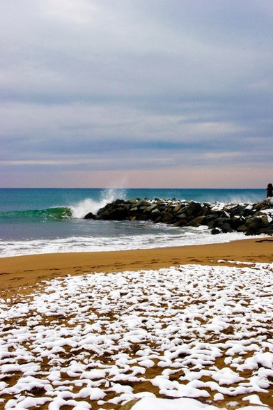 Photo au format vertical d’une plage de sable ocre et d’une digue de rochers noirs. Le ciel est couvert par une épaisse couche de nuages gris, tirant vers le rose au dessus de l’horizon. La mer a une jolie teinte bleu-vert foncé, et une vague se brise sur la digue. Au premier plan, une fine couche de neige recouvre le sable.