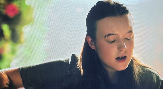 Bella Ramsey as Ellie strumming an acoustic guitar and singing. Her eyes are downward, looking at the cords. There's a pretty vine with a red flower behind her out of focus.
