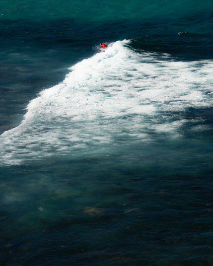 Aerial view of a surfer in a red shirt riding a white ocean wave. The deep blue and green sea contrasts with the cresting wave, creating dynamic movement across the water's surface.