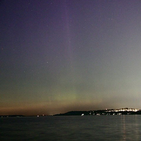 A night sky illuminated by stars, with a gradient of colors with shafts of purple to green, above a calm body of water. A distant shoreline is visible, dotted with lights.