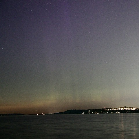 A serene night scene featuring a body of water under a starry sky, with soft hues of purple and green auroras visible on the horizon. A distant shoreline is lightly illuminated by city lights.