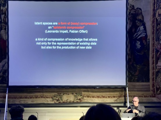 An individual speaks at a presentation, with a large screen behind displaying text about "latent spaces" and "epistemic compression." The setting features decorative walls and an audience in the foreground.