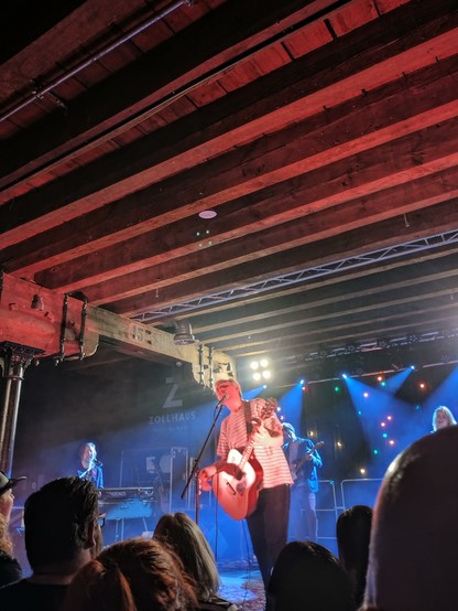 Foto von einer Bühne mit Musikern. Im Vordergrund steht ein Mann mit Gitarre und Mikro.