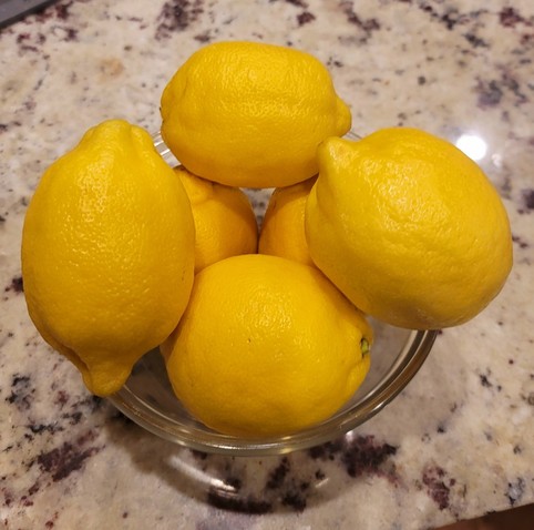 Clear bowl holding six lemons. Sitting on a purple, gray, and white granite counter top