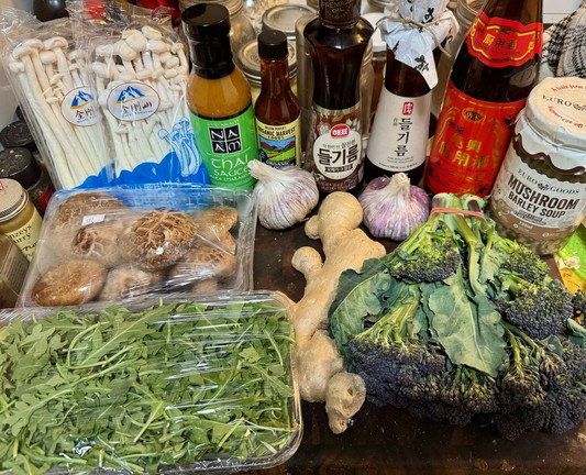 On a cutting board sits two packages of seafood mushrooms (white, with long narrow stalks) a package of shiitke mushrooms (brown),, a bottle of NAAM Thai sauce, a small bottle of chipotle-habaner sauce, two bottles of perilla oil, a larger bottle of Shaoxing cooking bine, a jar of mushroom barley soup, an enormous ginger root, two enormous heads of red garlic, a package of arugula and a bunch of red broccoli.